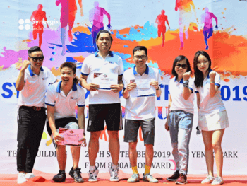 Group of people in white shirts stand on stage at a 2019 corporate event with colorful backdrop graphics.