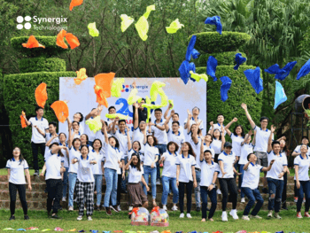 Large group of Synergix employees posing outdoors with colorful balloons and confetti celebrating together.