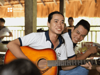 Two young men smile while playing an acoustic guitar together outdoors under a covered structure.