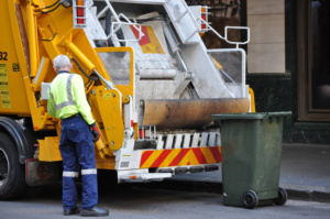 Waste management worker operates a yellow garbage truck with compactor mechanism during collection from a green bin.