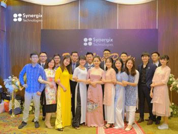 Large group of Synergix team members in business and traditional attire posing together at an indoor corporate event.